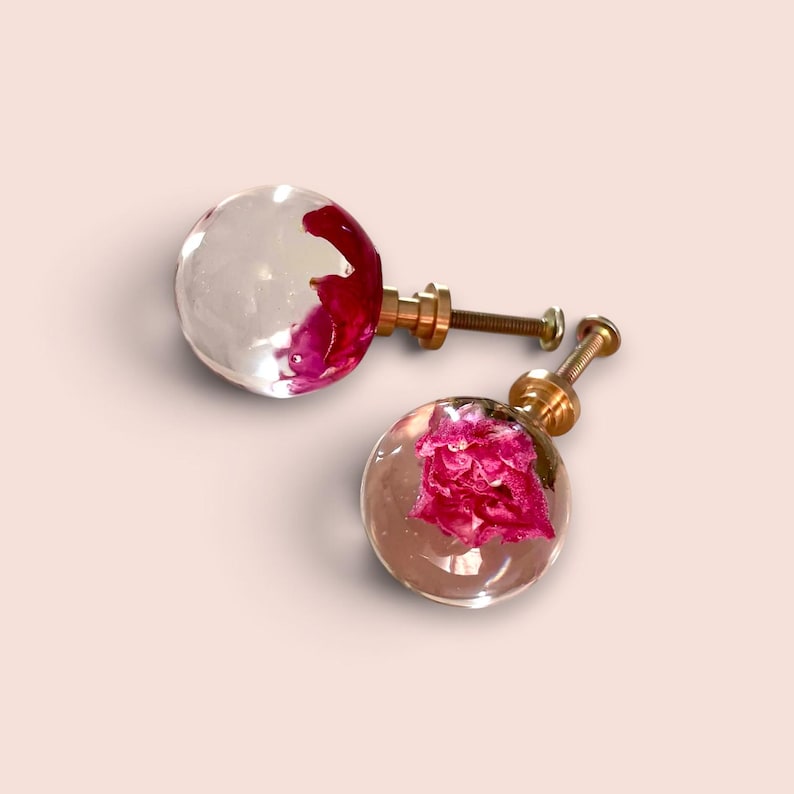 May include: Two spherical cabinet knobs featuring clear resin spheres with embedded bright pink flower petals. Each knob is attached to a gold-toned metal base with a threaded screw post extending from the back for installation on furniture.