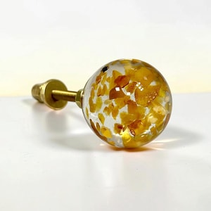 Puede incluir: Un pomo esf&eacute;rico para muebles con un cuerpo de resina transparente incrustado con numerosos fragmentos de piedra peque&ntilde;os e irregulares de color amarillo y &aacute;mbar, unido a un perno roscado de metal dorado.