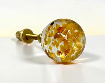 Op de afbeelding: Een bolvormige meubelknop met een heldere harsbehuizing waarin talloze kleine, onregelmatige gele en amberkleurige steenfragmenten zijn verwerkt, bevestigd aan een goudkleurige metalen bout met schroefdraad.