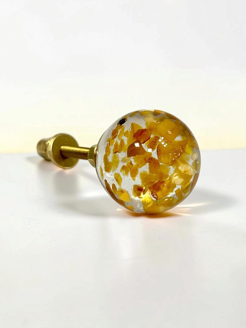 Puede incluir: Un pomo esf&eacute;rico para muebles con un cuerpo de resina transparente incrustado con numerosos fragmentos de piedra peque&ntilde;os e irregulares de color amarillo y &aacute;mbar, unido a un perno roscado de metal dorado.