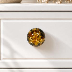 May include: A round cabinet knob featuring a clear resin dome filled with embedded amber, gold, and dark brown stone fragments. The knob is mounted on a smooth, white, rectangular drawer front with a recessed panel border design.