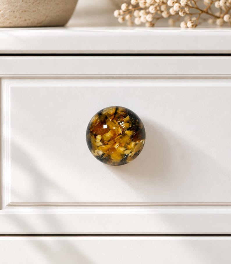 May include: A round cabinet knob featuring a clear resin dome filled with embedded amber, gold, and dark brown stone fragments. The knob is mounted on a smooth, white, rectangular drawer front with a recessed panel border design.
