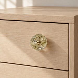May include: A faceted, clear resin drawer knob featuring embedded dried green foliage and gold-colored glitter particles. The knob is attached to a light-toned, wood-grain textured drawer front, showcasing a geometric, multi-sided shape.