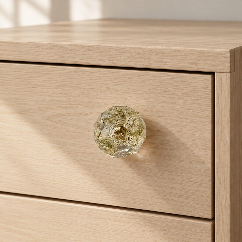 May include: A faceted, clear resin drawer knob featuring embedded dried green foliage and gold-colored glitter particles. The knob is attached to a light-toned, wood-grain textured drawer front, showcasing a geometric, multi-sided shape.