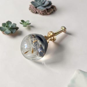 May include: A spherical cabinet knob featuring a clear resin dome encasing white dandelion seeds and dark blue accents, attached to a metallic gold-toned threaded bolt and a cylindrical base with a tiered design for secure mounting.
