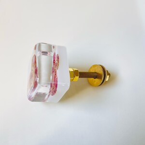 May include: A square cabinet knob made of clear resin with embedded pink floral elements and gold-colored metallic flakes. The knob features a gold-toned metal bolt, a nut, and a circular washer attached to the back for mounting purposes.