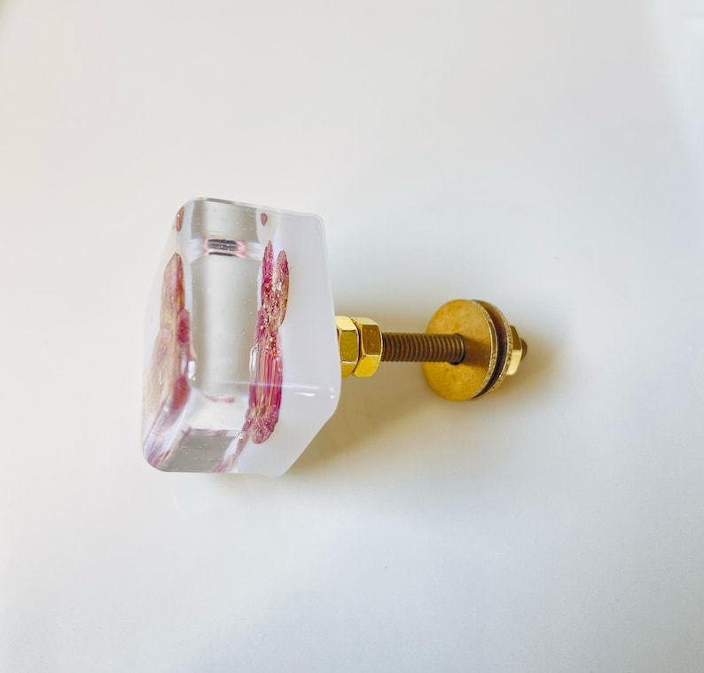 May include: A square cabinet knob made of clear resin with embedded pink floral elements and gold-colored metallic flakes. The knob features a gold-toned metal bolt, a nut, and a circular washer attached to the back for mounting purposes.