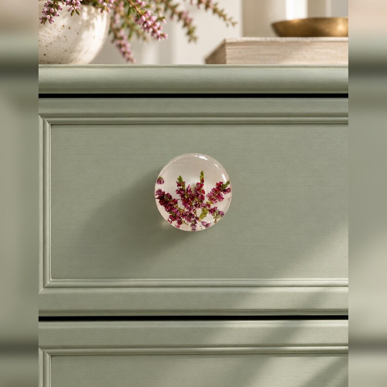 Puede incluir: Un tirador de caj&oacute;n redondo de resina transparente con flores de brezo moradas incrustadas. El tirador est&aacute; montado en un frente de caj&oacute;n de madera verde salvia con acabado mate y detalles de paneles horizontales.