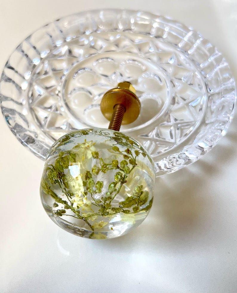 K&ouml;nnte beinhalten: Ein kugelf&ouml;rmiger M&ouml;belknopf aus klarem Harz mit eingebetteten gr&uuml;nen Trockenblumen. Der Knopf verf&uuml;gt &uuml;ber einen goldfarbenen Metallbolzen und eine kreisf&ouml;rmige Basisplatte mit einem facettierten, glasartigen Muster.