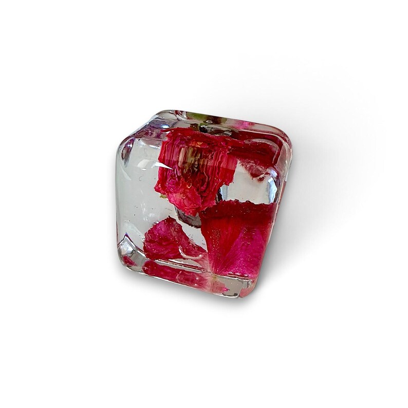 May include: A clear, cube-shaped resin paperweight containing vibrant, deep red rose petals suspended within the transparent material. The object features smooth, rounded edges and a polished finish, showcasing the natural texture of the petals.