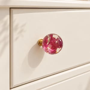 May include: A spherical drawer knob made of clear resin containing embedded magenta-colored dried flowers, attached to a brass-colored metal base, mounted on a smooth, off-white wooden drawer front with a rectangular frame detail.