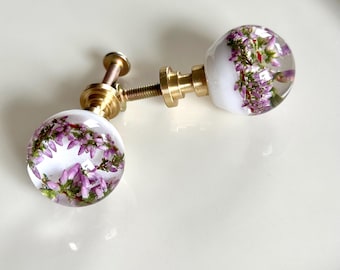Pode incluir: Dois puxadores esféricos para armários com esferas de resina transparente incrustadas com pequenas flores de urze roxas. Cada puxador tem uma base de metal em tom de latão e uma haste roscada para fixação.
