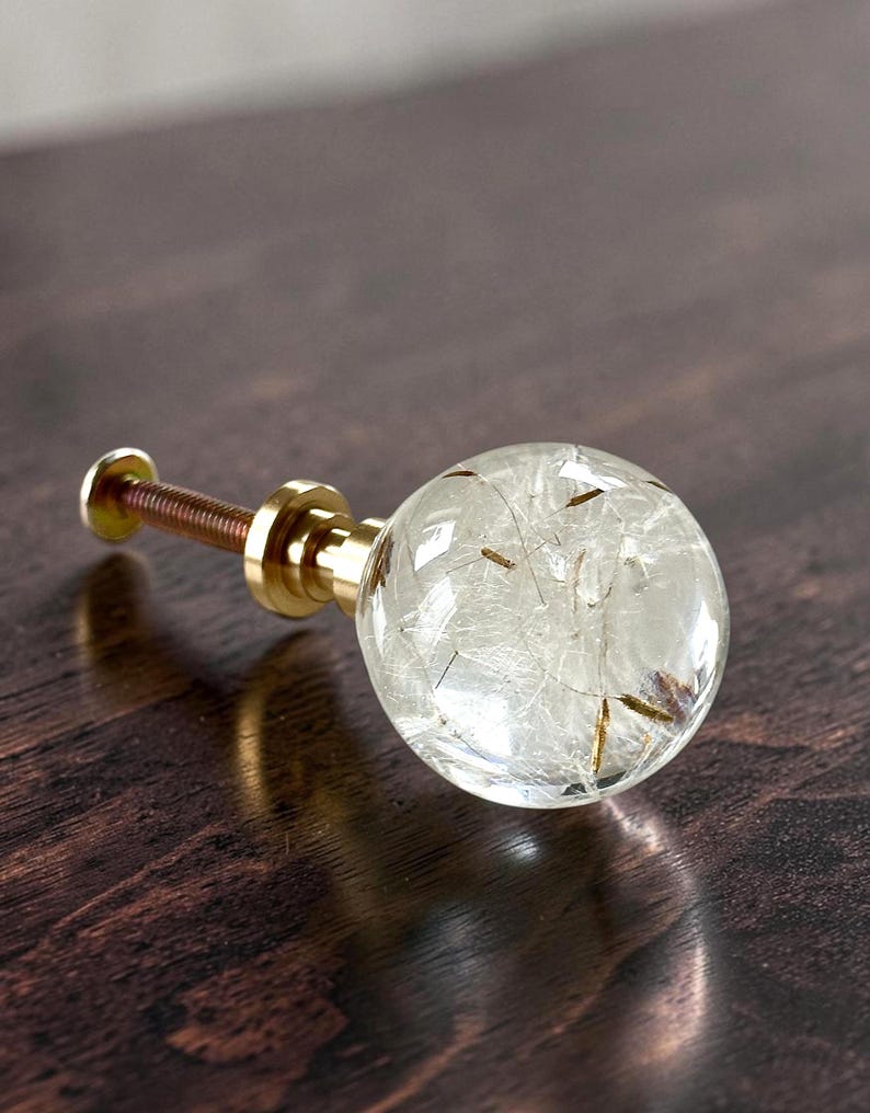 May include: A spherical cabinet knob featuring a clear resin orb containing delicate white dandelion seeds and brown stems. The knob is attached to a polished gold-tone metal base and a threaded metal screw for installation.