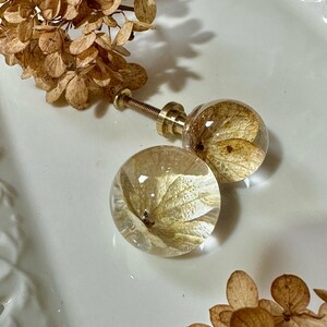 Puede incluir: Dos pomos esf&eacute;ricos para armarios hechos de resina transparente, cada uno con un p&eacute;talo de flor de hortensia seca de color marr&oacute;n claro. Los pomos cuentan con una base de metal dorado y un tornillo roscado, con un acabado liso y pulido.