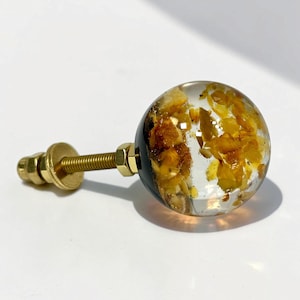 Peut inclure: Un bouton de meuble sphérique composé d'une résine transparente incrustée de flocons couleur ambre. Le bouton est fixé à une tige filetée en métal doré avec un écrou et une rondelle assortis, formant une pièce de quincaillerie fonctionnelle.