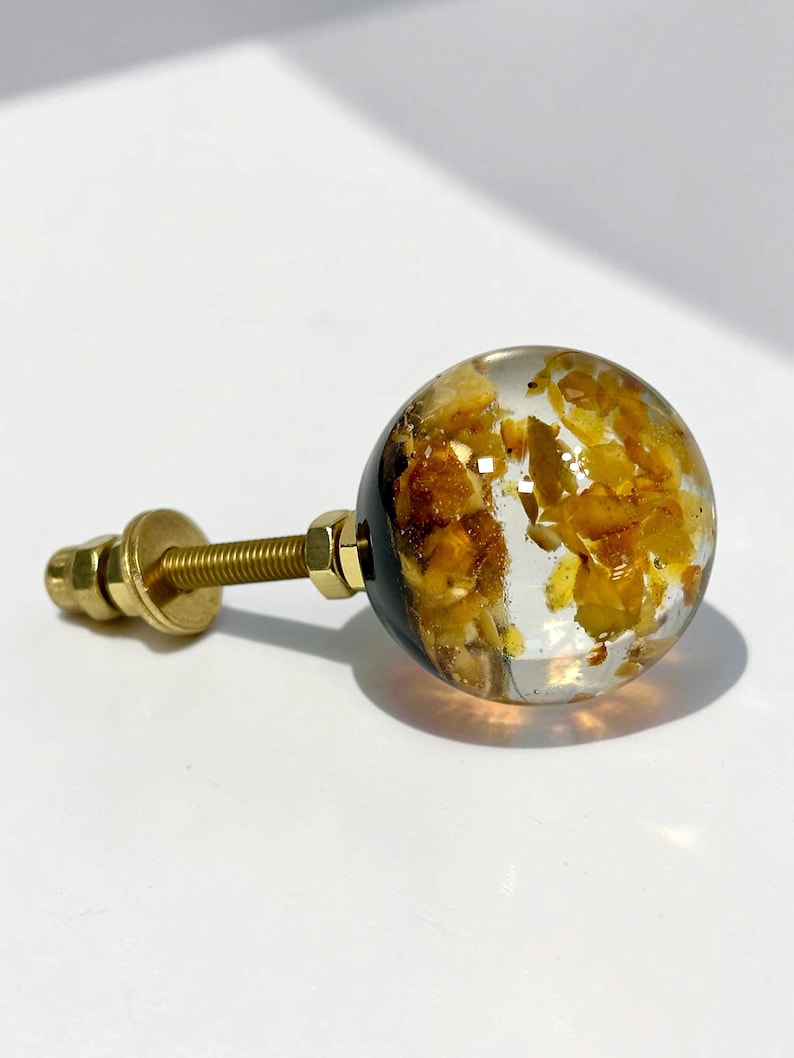 May include: A spherical cabinet knob featuring a clear resin body embedded with small, irregular amber-colored fragments. The knob is attached to a gold-toned metal bolt with a matching nut and washer, providing a secure mounting mechanism.