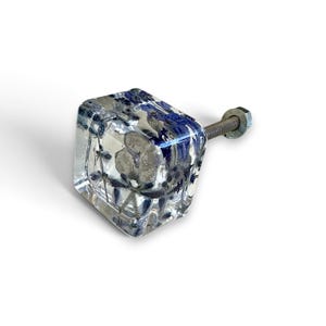 Op de afbeelding: Een vierkante meubelknop van heldere hars met ingebedde gedroogde blauwe bloemen en kleine grijze steentjes. De knop is voorzien van een metalen draadeind aan de achterkant, compleet met een kleine metalen moer bevestigd aan de schroef.