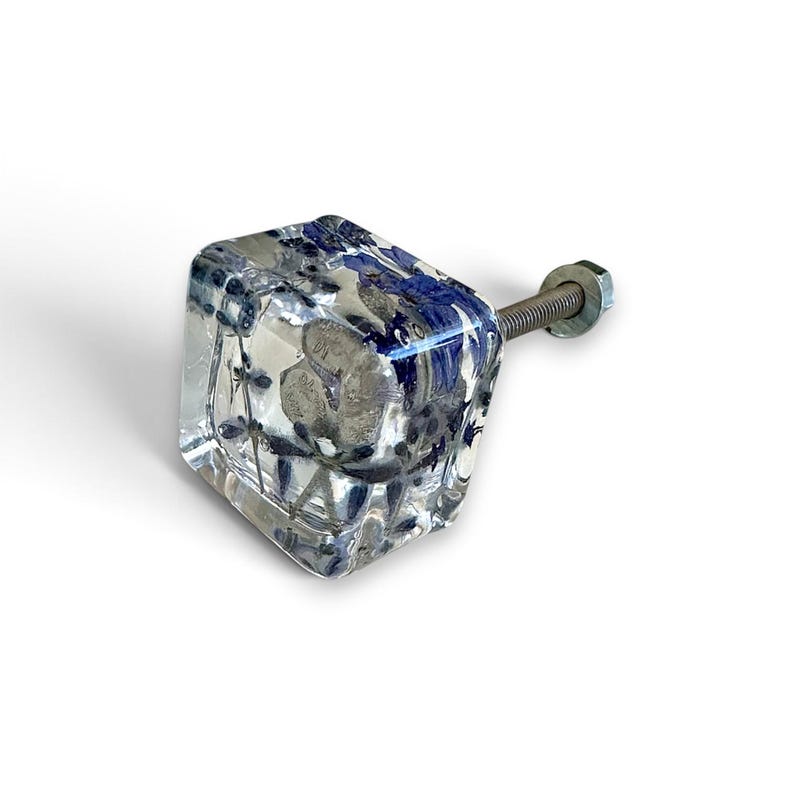 Op de afbeelding: Een vierkante meubelknop van heldere hars met ingebedde gedroogde blauwe bloemen en kleine grijze steentjes. De knop is voorzien van een metalen draadeind aan de achterkant, compleet met een kleine metalen moer bevestigd aan de schroef.
