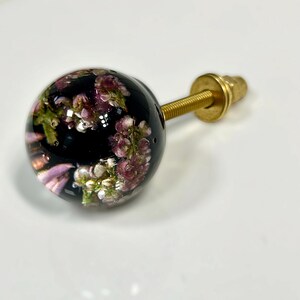 Pode incluir: Um puxador esf&eacute;rico para m&oacute;veis com acabamento em resina transparente contendo flores de urze roxas e brancas. O puxador est&aacute; fixado a um parafuso de metal dourado com uma anilha e porca correspondentes para a montagem.