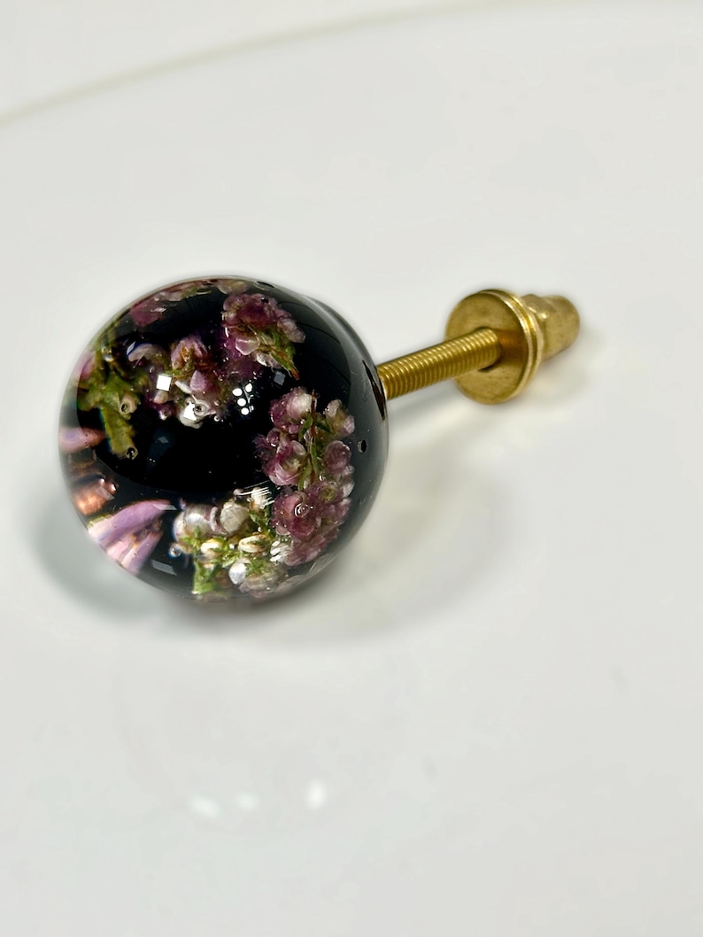 Pode incluir: Um puxador esf&eacute;rico para m&oacute;veis com acabamento em resina transparente contendo flores de urze roxas e brancas. O puxador est&aacute; fixado a um parafuso de metal dourado com uma anilha e porca correspondentes para a montagem.