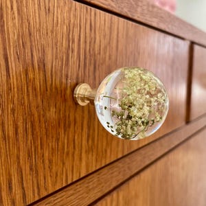 Pode incluir: Um puxador de gaveta esf&eacute;rico de resina transparente com flores brancas secas incrustadas no interior. O puxador est&aacute; fixado a uma base met&aacute;lica de tom lat&atilde;o, montada numa frente de gaveta de madeira com veios vis&iacute;veis e linhas horizontais.
