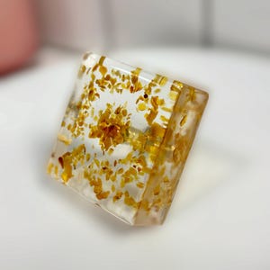 Op de afbeelding: Een vierkante ring met een heldere, transparante harszetting en ringband. De hars is ingelegd met talloze kleine, onregelmatige goudgele vlokken die verspreid zijn over het vierkante oppervlak en de band van de ring.