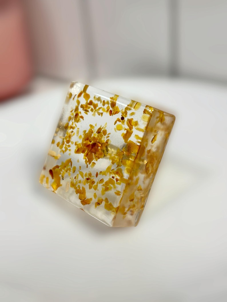 Op de afbeelding: Een vierkante ring met een heldere, transparante harszetting en ringband. De hars is ingelegd met talloze kleine, onregelmatige goudgele vlokken die verspreid zijn over het vierkante oppervlak en de band van de ring.