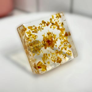 Op de afbeelding: Een helder, vierkant blok van hars met kleine, verspreide gele en amberkleurige vlokken. Het object heeft een gladde, glanzende afwerking met een transparant uiterlijk, waarbij de zwevende gouden deeltjes in het interieur zichtbaar zijn.