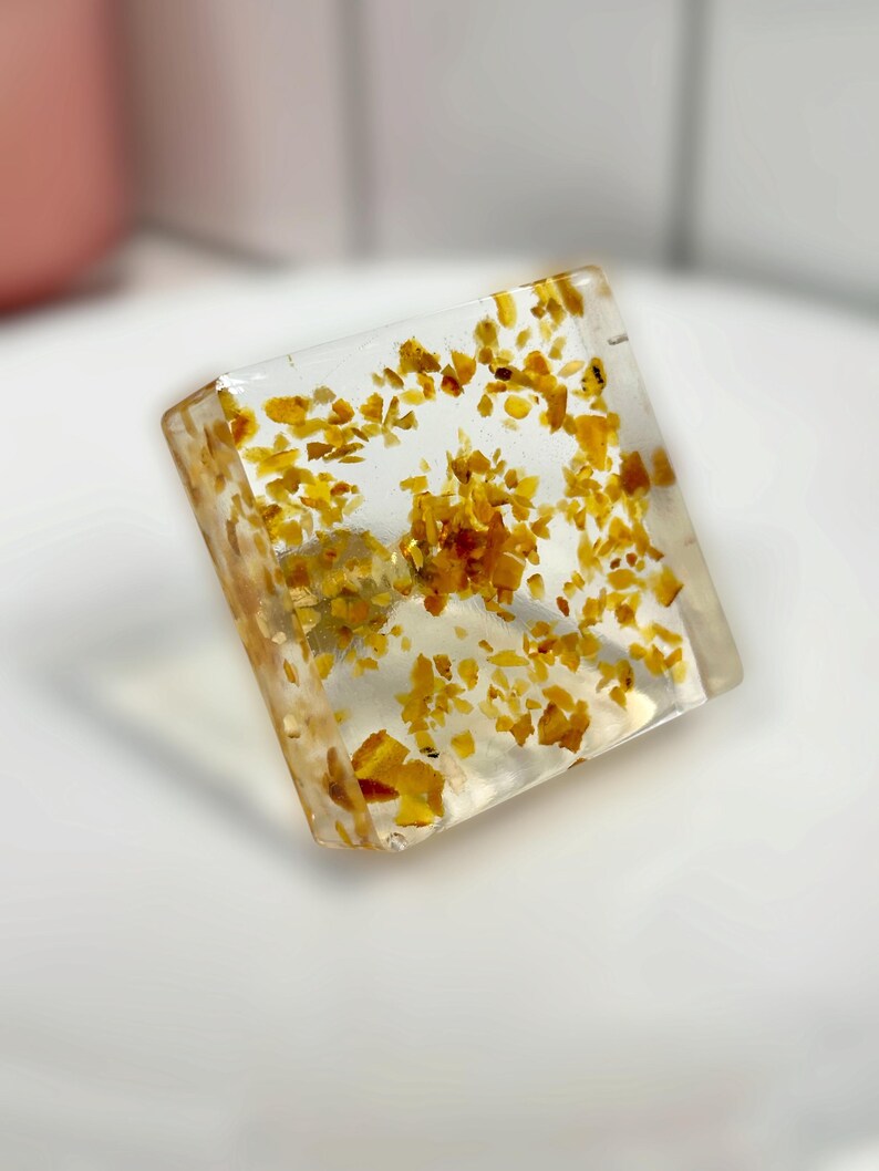 Op de afbeelding: Een helder, vierkant blok van hars met kleine, verspreide gele en amberkleurige vlokken. Het object heeft een gladde, glanzende afwerking met een transparant uiterlijk, waarbij de zwevende gouden deeltjes in het interieur zichtbaar zijn.