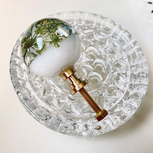 May include: A spherical cabinet knob featuring a clear resin top embedded with delicate green pressed flowers over a solid white base. The knob includes a metallic gold-toned threaded bolt and hardware for attachment to furniture or cabinetry.