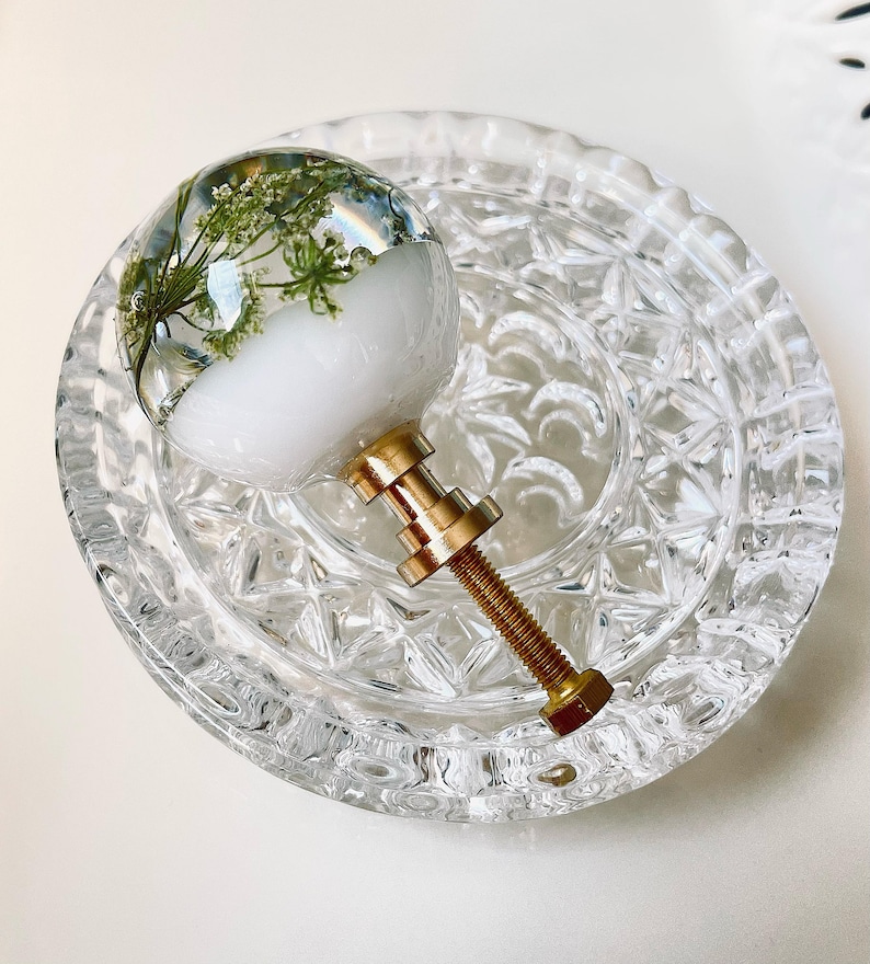 May include: A spherical cabinet knob featuring a clear resin top embedded with delicate green pressed flowers over a solid white base. The knob includes a metallic gold-toned threaded bolt and hardware for attachment to furniture or cabinetry.