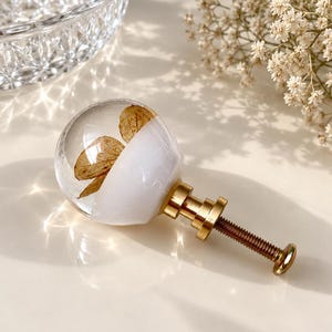 May include: A spherical cabinet knob featuring a clear resin top half containing a dried brown flower petal and an opaque white bottom half. The knob is attached to a metallic gold-toned base with a threaded screw extending from the back.