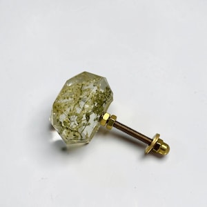 Pode incluir: Um puxador de m&oacute;vel hexagonal, transparente e facetado, contendo flores brancas secas embutidas. O puxador possui um parafuso, uma anilha e uma porca de metal dourado na parte posterior, formando uma pe&ccedil;a de ferragem funcional.