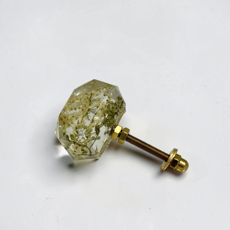 Pode incluir: Um puxador de m&oacute;vel hexagonal, transparente e facetado, contendo flores brancas secas embutidas. O puxador possui um parafuso, uma anilha e uma porca de metal dourado na parte posterior, formando uma pe&ccedil;a de ferragem funcional.
