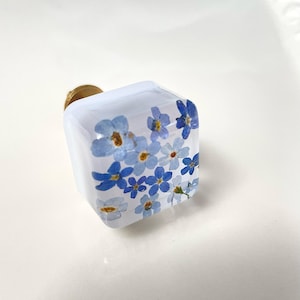 Puede incluir: Un tirador de mueble cuadrado con un acabado transparente y brillante que contiene flores de nomeolvides azules prensadas. El tirador tiene una forma c&uacute;bica de bordes redondeados e incluye una base circular de metal dorado para su montaje.