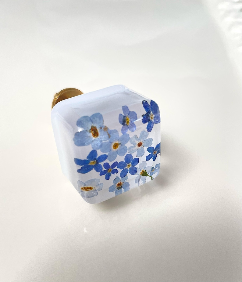 Puede incluir: Un tirador de mueble cuadrado con un acabado transparente y brillante que contiene flores de nomeolvides azules prensadas. El tirador tiene una forma c&uacute;bica de bordes redondeados e incluye una base circular de metal dorado para su montaje.