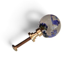 May include: A spherical cabinet knob made of clear resin containing embedded blue flowers. The knob features a metallic gold-toned base and a threaded metal bolt extending from the back for installation on furniture or cabinetry doors.
