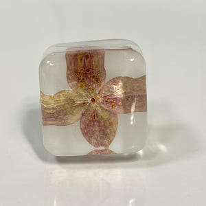 May include: A clear, square-shaped resin block containing a single, pressed pink and yellow hydrangea flower. The transparent material has rounded edges and corners, showcasing the delicate, dried petals of the flower suspended within the center.