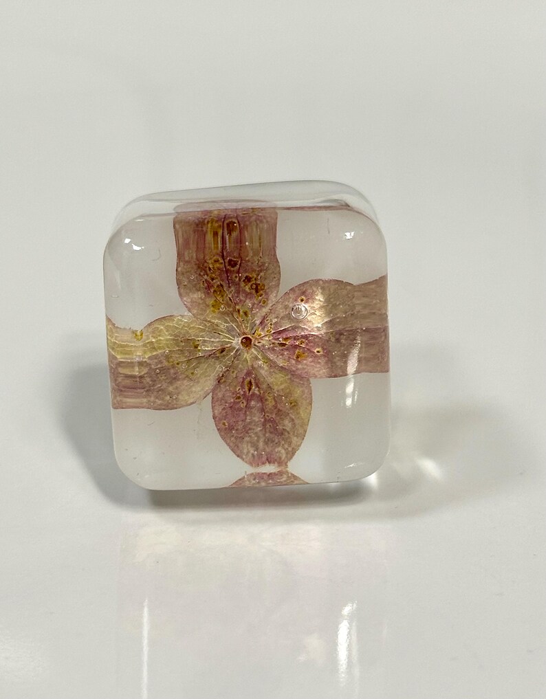 May include: A clear, square-shaped resin block containing a single, pressed pink and yellow hydrangea flower. The transparent material has rounded edges and corners, showcasing the delicate, dried petals of the flower suspended within the center.