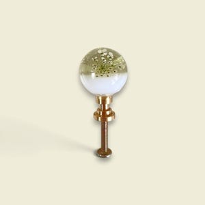 May include: A spherical cabinet knob featuring a clear resin top half embedded with small white pressed flowers and a solid white opaque bottom half. The knob is attached to a metallic gold-toned threaded bolt and mounting hardware base.