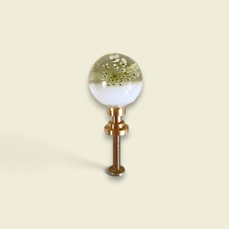 May include: A spherical cabinet knob featuring a clear resin top half embedded with small white pressed flowers and a solid white opaque bottom half. The knob is attached to a metallic gold-toned threaded bolt and mounting hardware base.