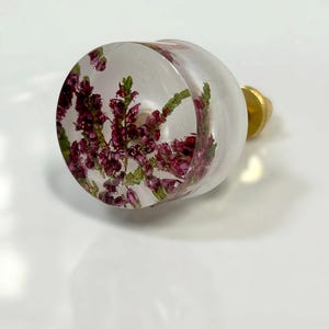 May include: A round cabinet knob made of clear resin with embedded purple heather flowers and green stems. The knob features a flat circular face and a gold-toned metal base attachment extending from the back for mounting purposes.