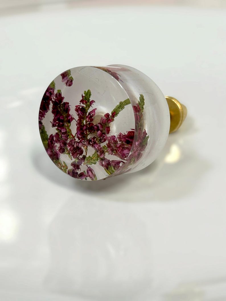 Puede incluir: Un pomo redondo para muebles hecho de resina transparente con flores de brezo moradas y tallos verdes incrustados. El pomo cuenta con una base de metal dorado y un herraje de fijaci&oacute;n en la parte posterior para su uso funcional.