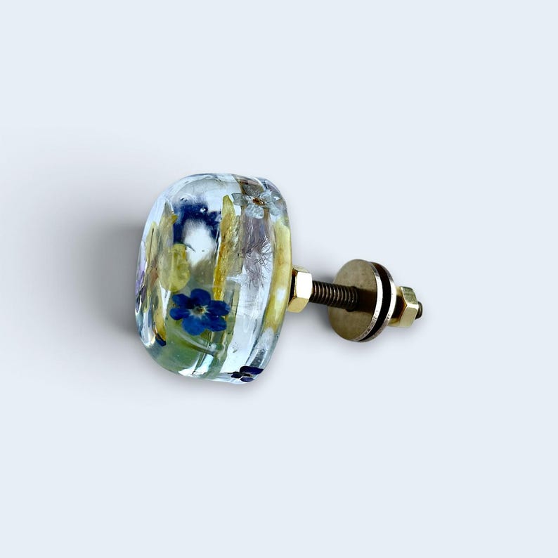 May include: A clear, rounded cabinet knob featuring embedded dried blue flowers and yellow botanical elements. The knob is attached to a metallic threaded bolt with a brass-colored nut and washer, providing a functional hardware component.