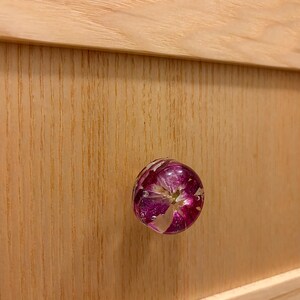 May include: A round cabinet knob featuring a clear resin dome that encapsulates a vibrant magenta and white flower. The knob is attached to a light-colored wooden drawer front with a visible vertical wood grain pattern and horizontal trim.