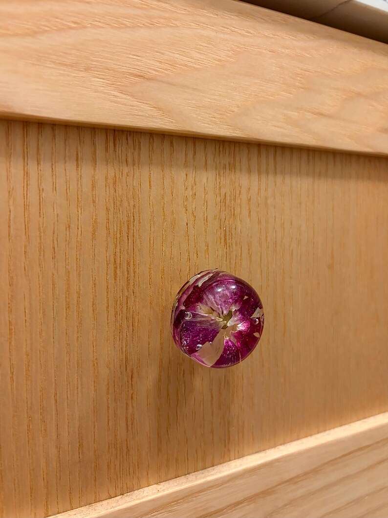 May include: A round cabinet knob featuring a clear resin dome that encapsulates a vibrant magenta and white flower. The knob is attached to a light-colored wooden drawer front with a visible vertical wood grain pattern and horizontal trim.