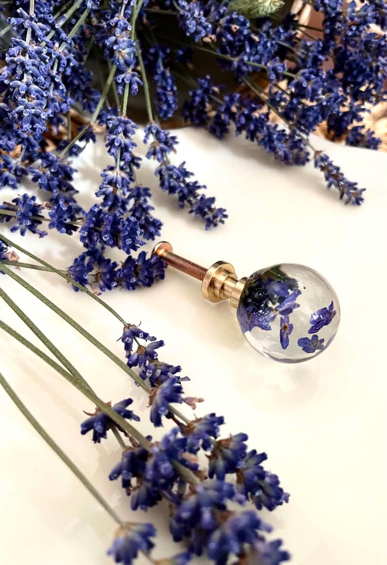 May include: A spherical cabinet knob made of clear resin containing dried purple lavender flowers. The knob features a gold-toned metal base and a threaded screw attachment, providing a functional and decorative hardware piece for furniture.