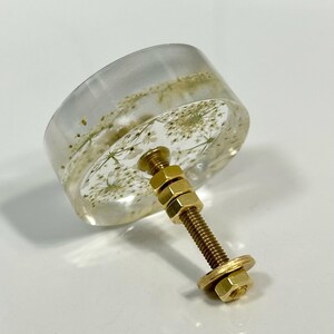 Puede incluir: Un pomo redondo para mueble hecho de resina transparente con flores blancas secas incrustadas. El pomo est&aacute; unido a un perno de metal dorado con dos tuercas hexagonales ajustables y una arandela plana en el extremo.