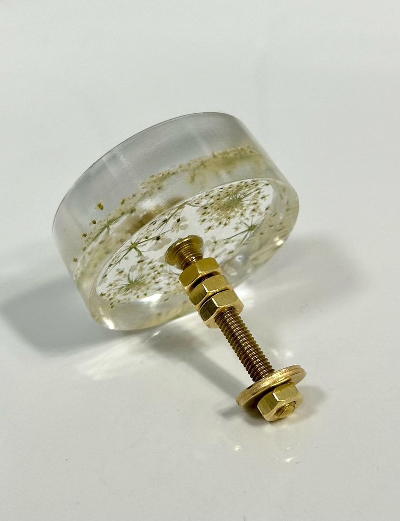Puede incluir: Un pomo redondo para mueble hecho de resina transparente con flores blancas secas incrustadas. El pomo est&aacute; unido a un perno de metal dorado con dos tuercas hexagonales ajustables y una arandela plana en el extremo.