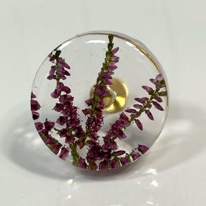 Può includere: Un pomello circolare in resina trasparente con fiori di erica viola incorporati. Il pomello ha una finitura liscia e trasparente e include una base di montaggio in metallo dorato al centro, progettata per mobili o armadietti.